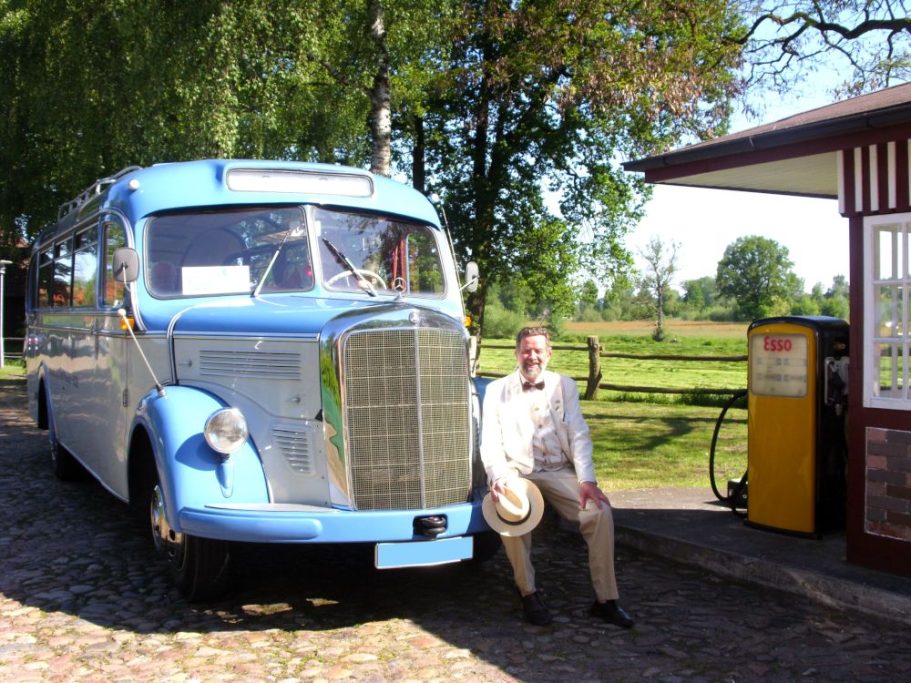 Der Busfahrer sitzt auf der Stoßstange eines blauen Oldtimerbus Mercedes